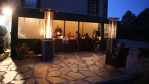 Une terrasse extérieure avec un sol en pierre, des tables, des chaises et un bâtiment doté de grandes fenêtres en arrière-plan.