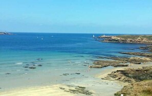 Una playa costera con aguas cristalinas, un litoral rocoso y un faro a lo lejos.