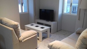 Ein Wohnzimmer mit einem beigefarbenen Sofa, einem weißen Couchtisch und einem Flachbildfernseher auf einem Standfuß.