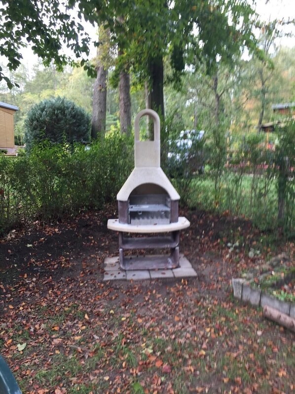 A stone outdoor grill with a chimney, surrounded by a garden with trees and bushes.