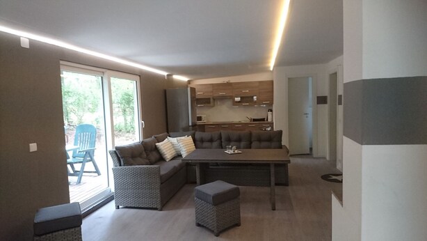 Una sala moderna con un sofá seccional, una mesa de comedor y un pequeño reposapiés. Hay una puerta corrediza de vidrio que da a un espacio exterior con una silla azul. Se ve la cocina al fondo, con armarios de madera y un microondas.