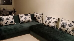 Un sofá seccional verde con cojines de estampado floral, una mesa de comedor con sillas y una ventana con persianas.