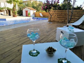 A poolside area with wooden decking, two lounge chairs, and a table with a bowl of olives and glasses of water.