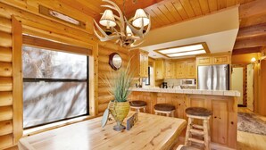A log cabin interior with a wooden dining table, bar stools, a kitchen with stainless steel appliances, and a large window. - Knotty AND Nice #177 (Big Bear Lake)