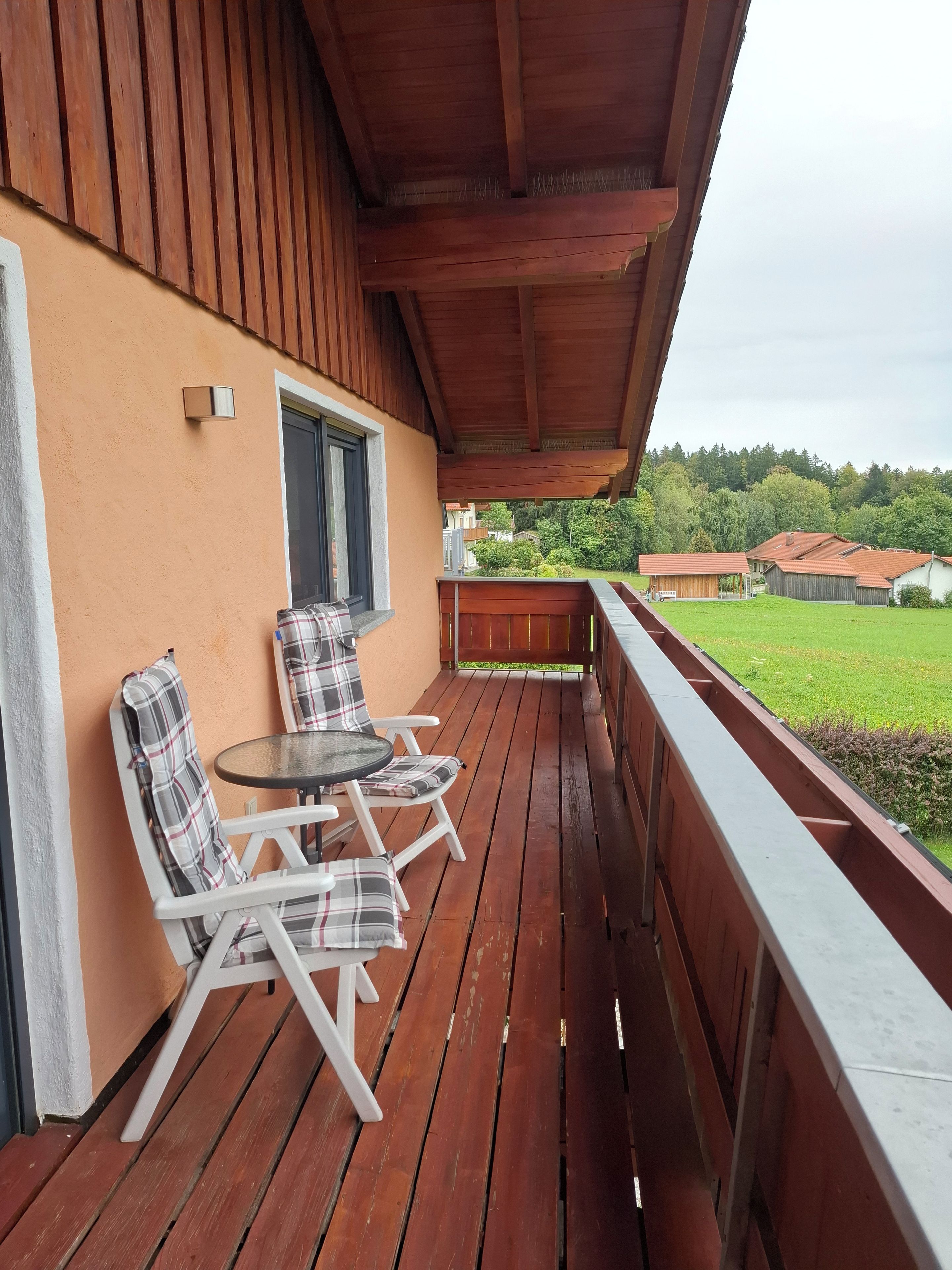 Eine Holzterrasse mit Tisch und Stühlen, ein Fenster und Blick auf eine grüne Landschaft.