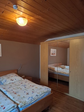 Ein Zimmer mit Holzboden, einem Bett mit floralem Bettbezug, einem Nachttisch, einem Spiegel und einer Deckenleuchte.