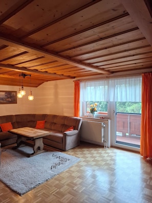 Ein gemütliches Wohnzimmer mit Holzboden, einem karierten Sofa, einem Tisch und Blick durch ein Fenster mit orangefarbenen Vorhängen.
