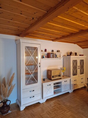 A room with wooden ceiling beams, a white cabinet with glass doors, and a shelf with various items. - 1A Apartment TOP (Klingenbrunn)