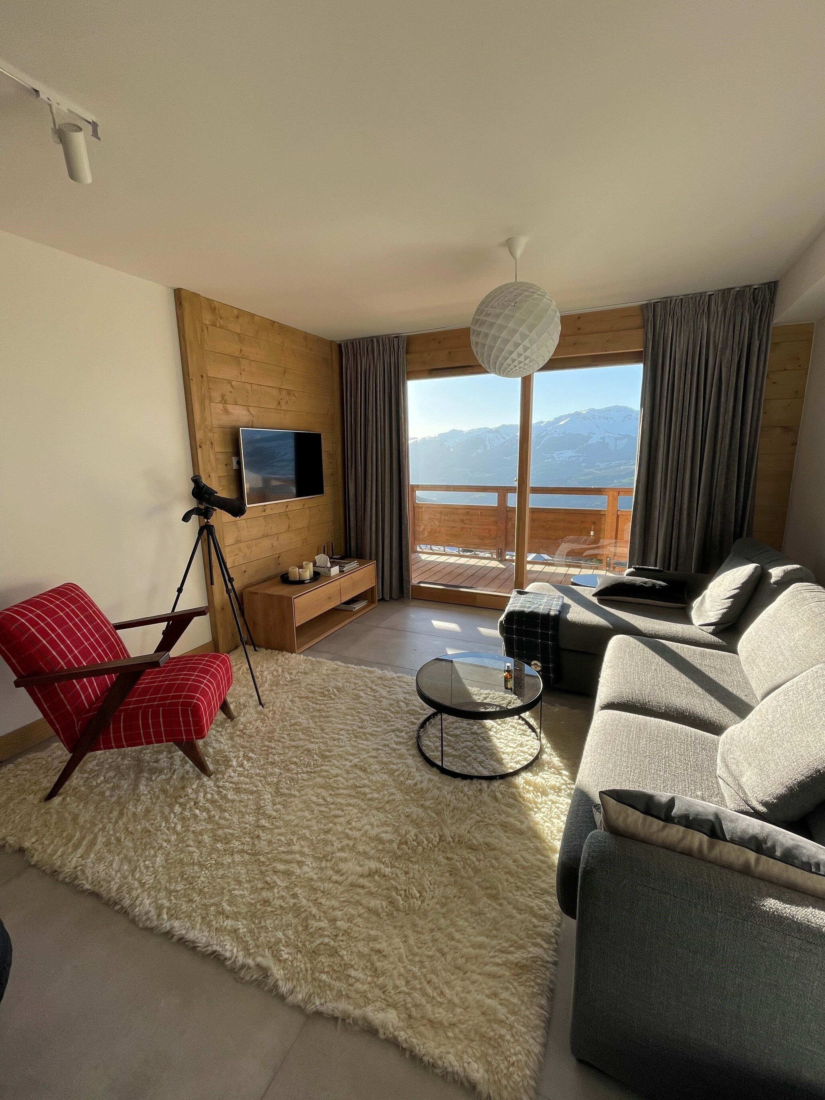 A modern living room with a large window offering a mountain view, a red chair, a grey sofa, and a small round table.