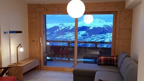 A modern living room with a large window offering a mountain view, a grey sofa, a red chair, and a wooden bench. - Interior comfort and divine sensations outside. New MGM apartment. (Peisey-Vallandry)
