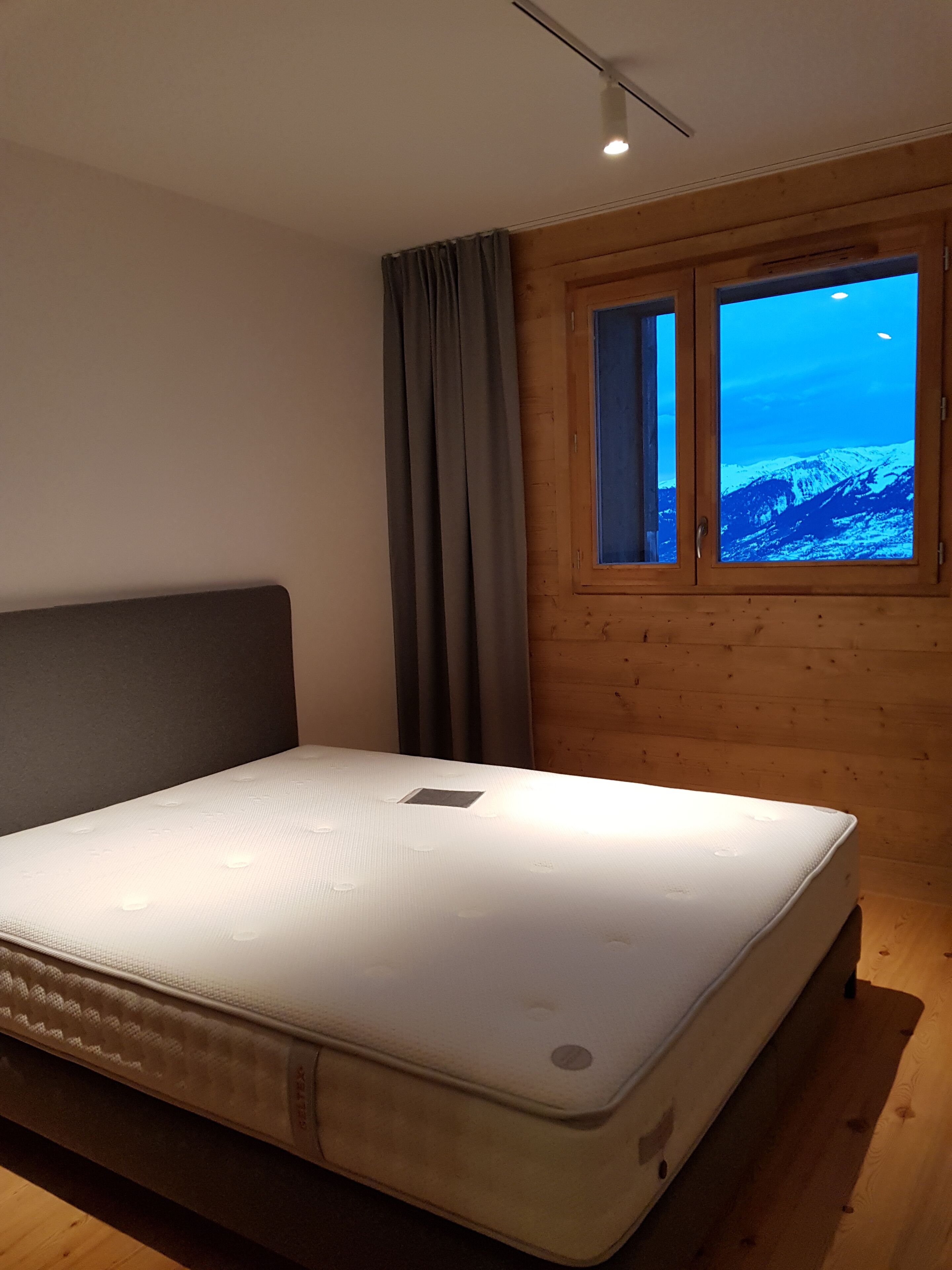 A bedroom with a single mattress, wooden walls, a window with a view of a snowy mountain, and a curtain.
