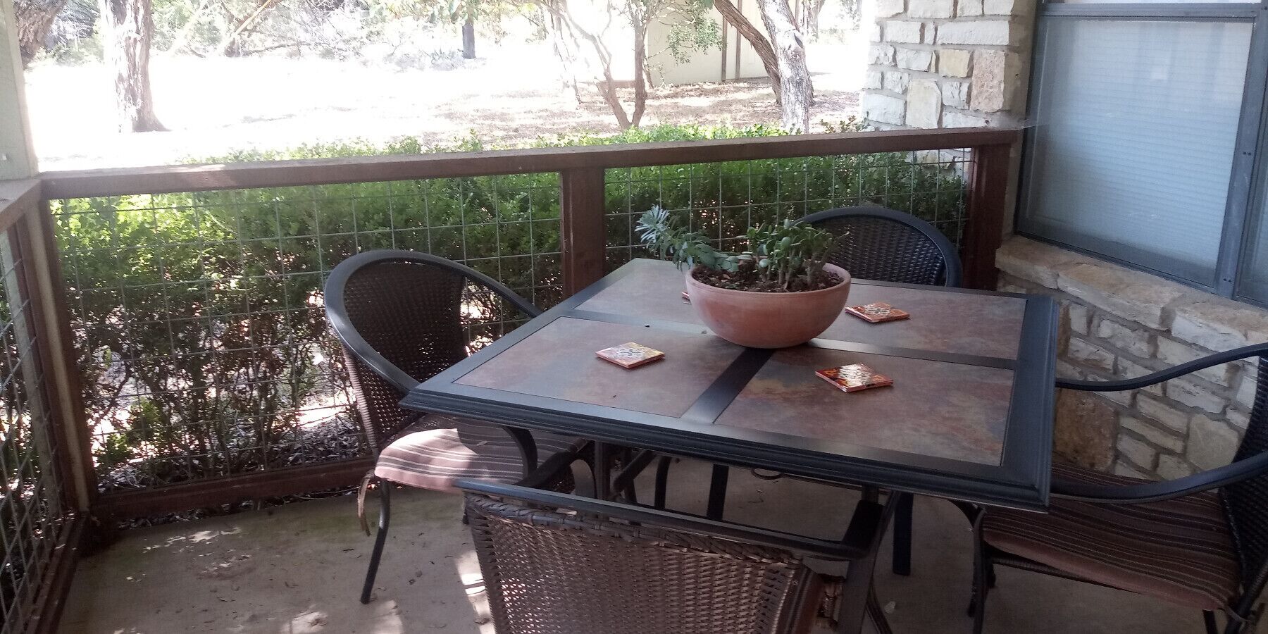 A small outdoor patio with a table and chairs, a potted plant, and a view of greenery.