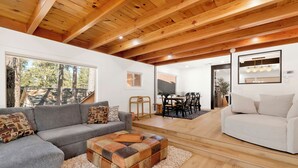 A spacious living area with a grey sectional sofa, a white sofa, and a wooden coffee table. There is a dining area with a table and chairs, and a large window providing a view of trees and a building. - Vista Pines #234 (Big Bear Lake)