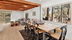 A modern dining area with a wooden table and chairs, a sofa, and a view of trees through large windows. - Vista Pines #234 (Big Bear Lake)