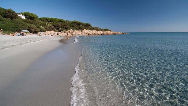 Ein Strand mit klarem Wasser, sandigem Ufer und einem felsigen Vorsprung mit Vegetation.