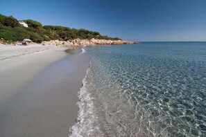 Ein Strand mit klarem Wasser, sandigem Ufer und einem felsigen Vorsprung mit Vegetation.
