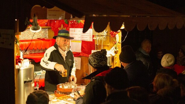Un puesto de comida con un cartel de menú, una persona sirviendo cerveza y una multitud de gente.