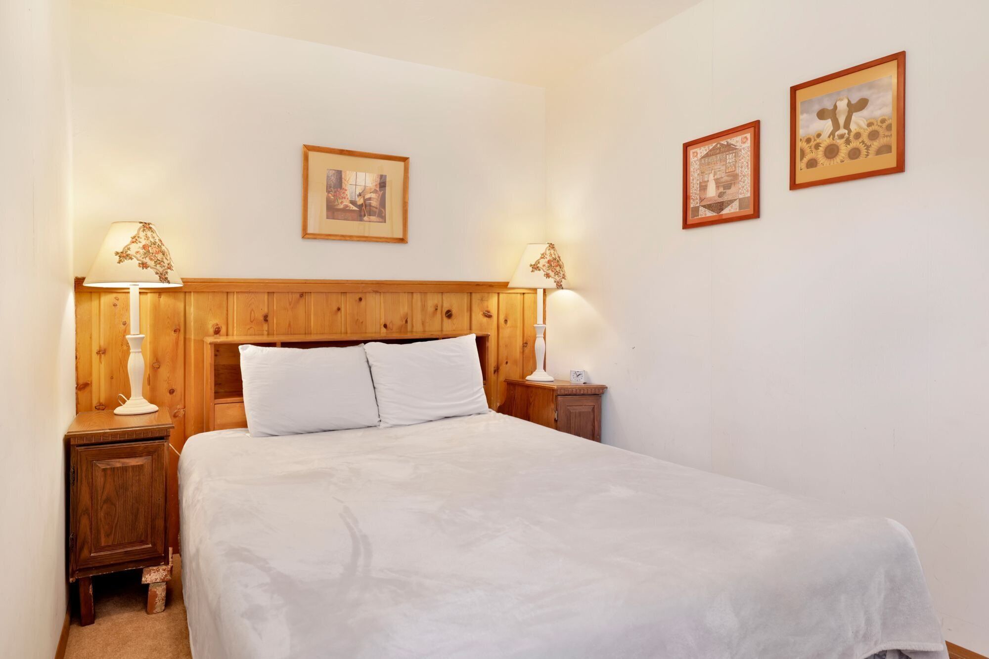 A neatly made bed with white linens in a room with wooden paneling on the walls and a wooden headboard. There are two bedside tables with lamps and framed pictures on the wall.