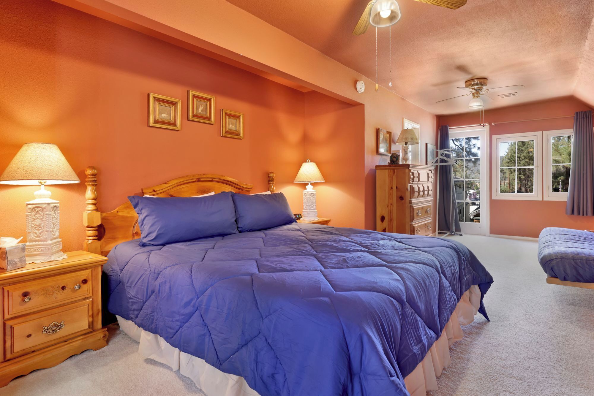 A bedroom with a large bed, two bedside tables with lamps, a wooden dresser, and a window with a view of trees.