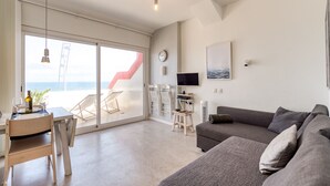 Una sala de estar moderna con un sofá gris, una mesa de comedor con sillas, un televisor y un ventanal con vistas al mar.