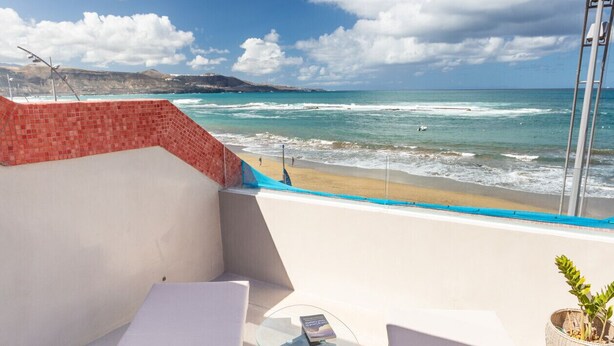Un balcón con dos sillones de mimbre y una pequeña mesa, con vistas a una playa, olas y cielo despejado.