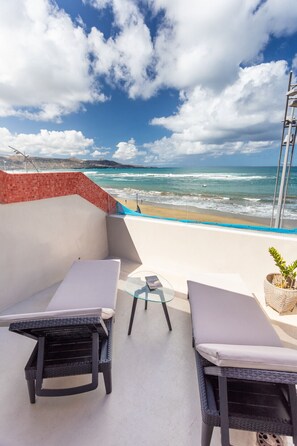 A balcony with two wicker chairs and a small table, overlooking a beach with waves and a clear sky.