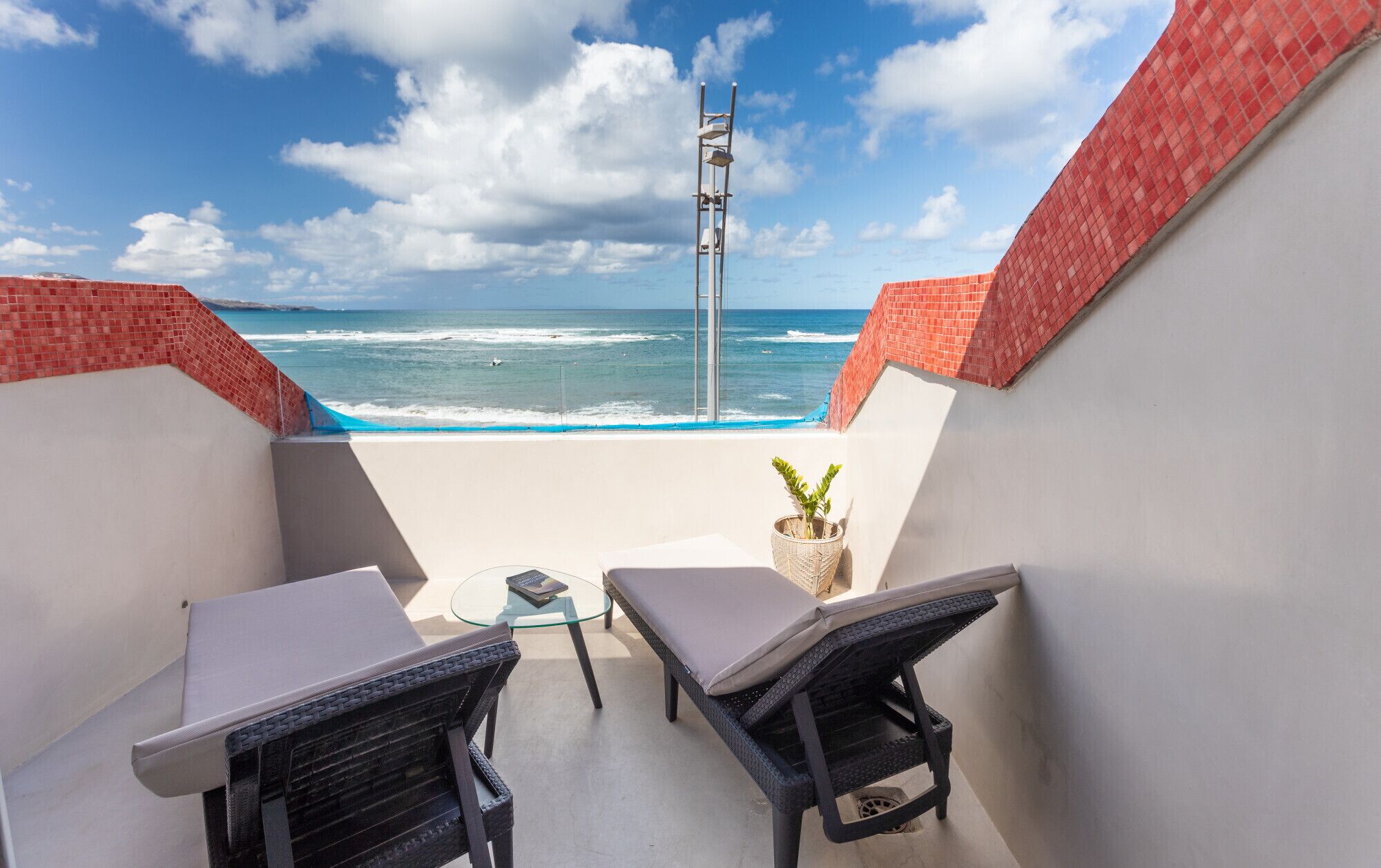 A balcony with two wicker chairs, a small table, and a potted plant. The balcony overlooks the ocean with waves crashing onto the shore.