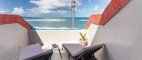 A balcony with two wicker chairs, a small table, and a potted plant. The balcony overlooks the ocean with waves crashing onto the shore.
