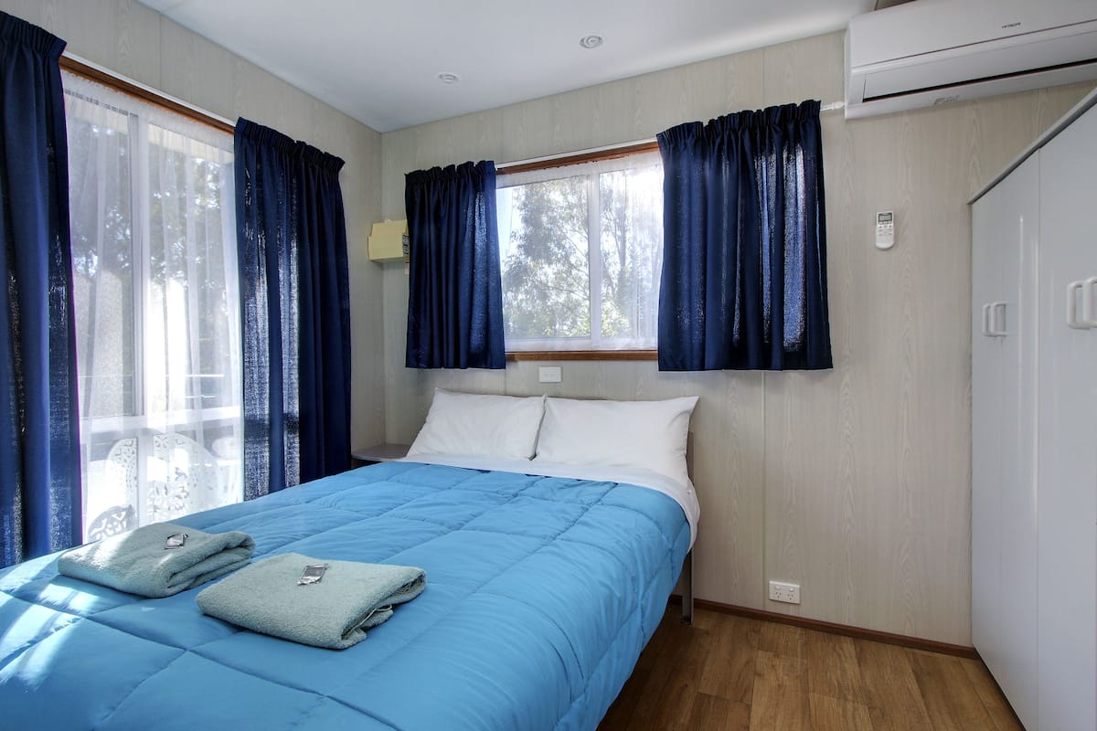 A bedroom with a bed, two towels, a window with curtains, an air conditioner, and a wardrobe.