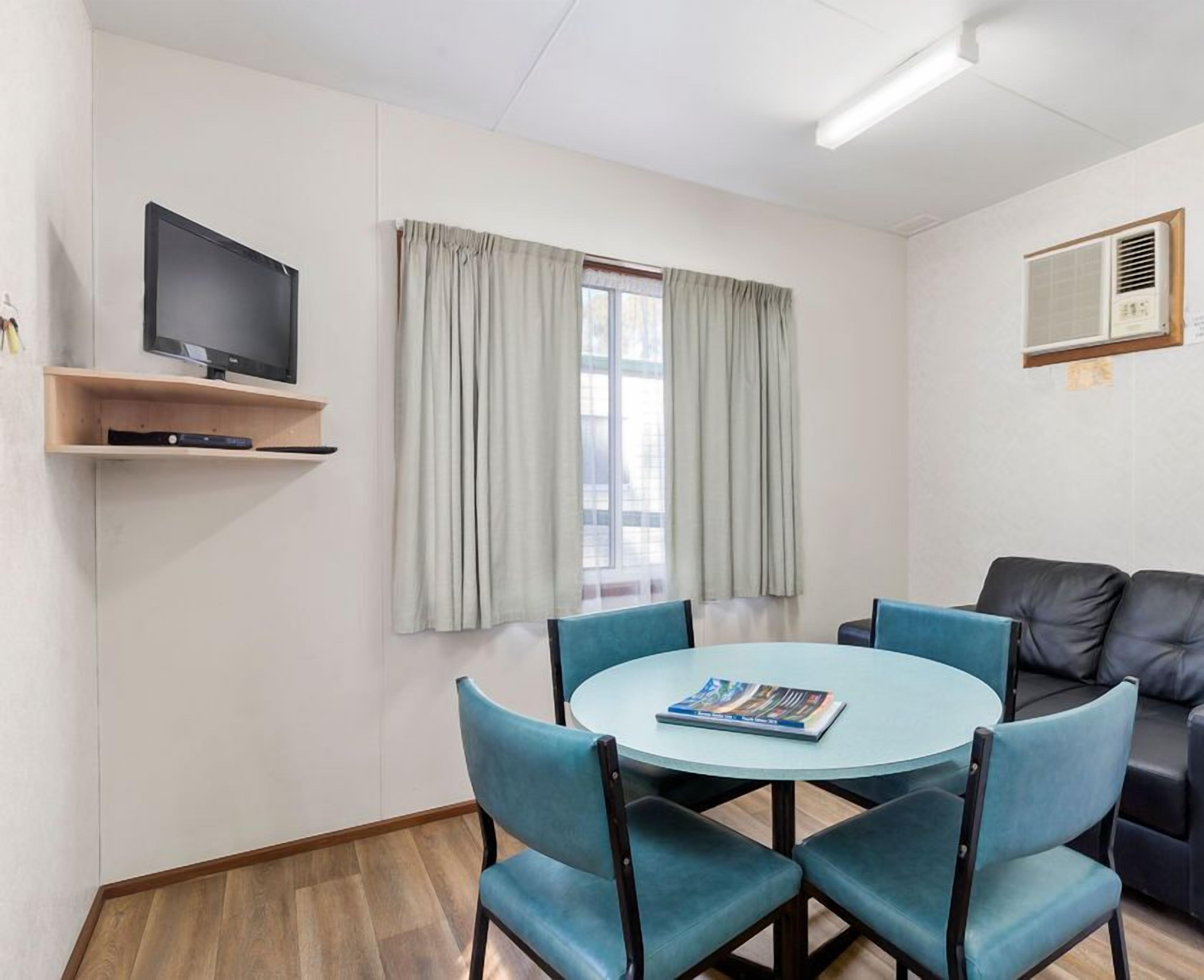 Una habitación con una mesa redonda, cuatro sillas, un televisor en un estante, una ventana con cortinas, un sofá y un equipo de aire acondicionado.