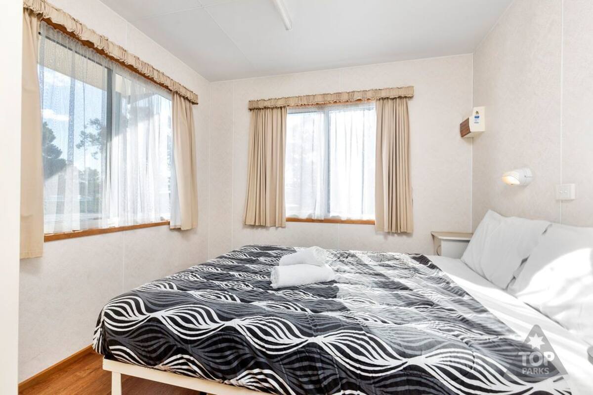 A bedroom with a bed, two windows with curtains, a bedside table, and a wall-mounted light.