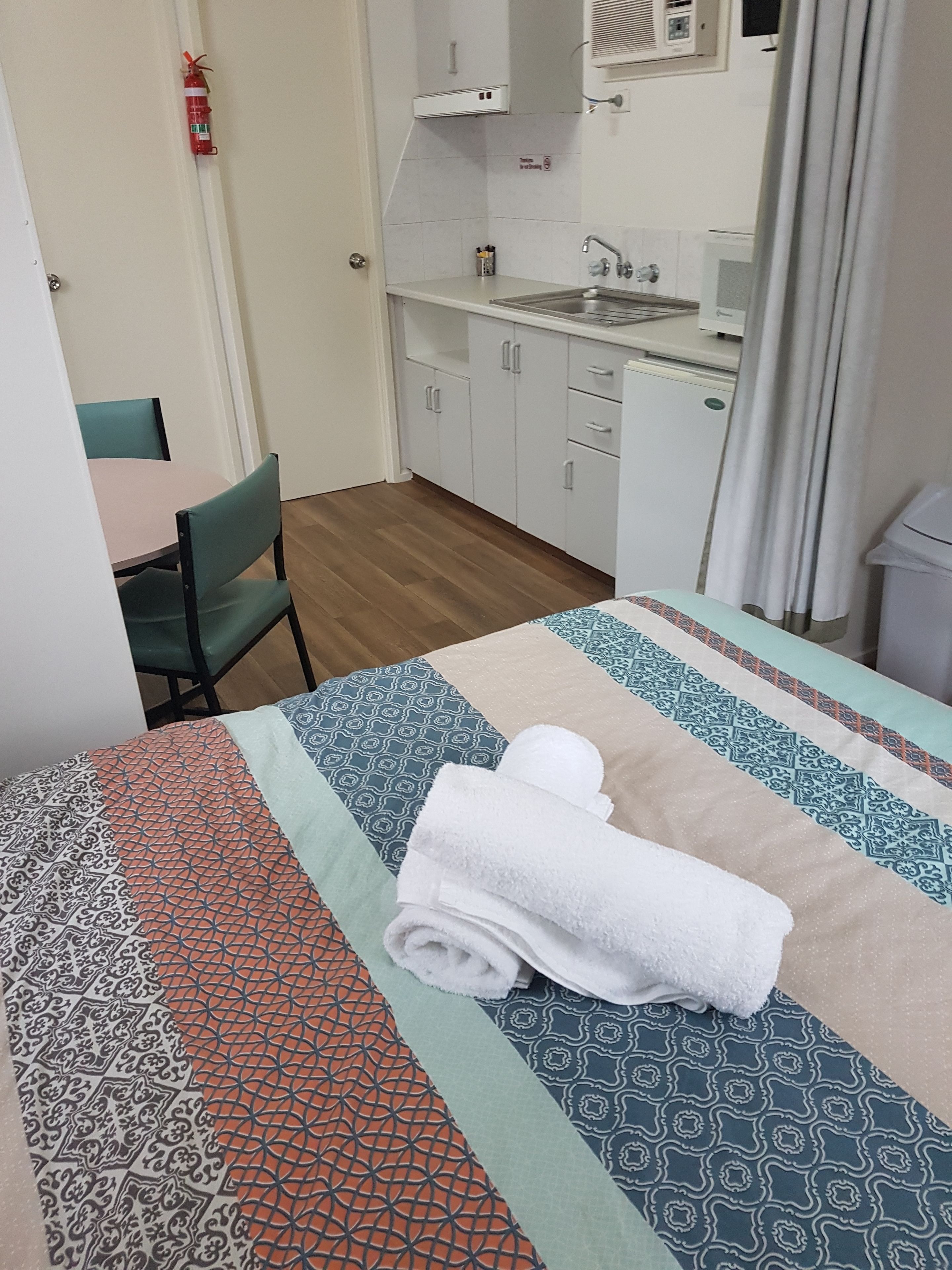 A hotel room with a bed, a small desk, a chair, a kitchenette, and a fire extinguisher.