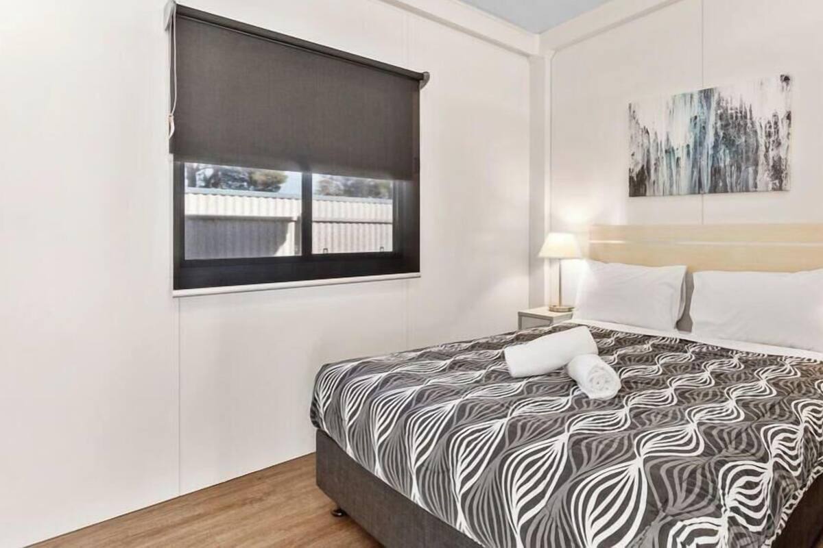 A hotel room with a bed, a window with a blind, a nightstand with a lamp, and a painting on the wall.