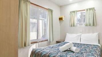 Una habitación de hotel con una cama, dos ventanas con cortinas, una mesita de noche y una lámpara fijada a la pared.