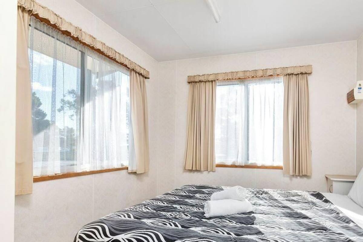 A hotel room with a large bed, two windows with curtains, a bedside table, and a wall-mounted light.