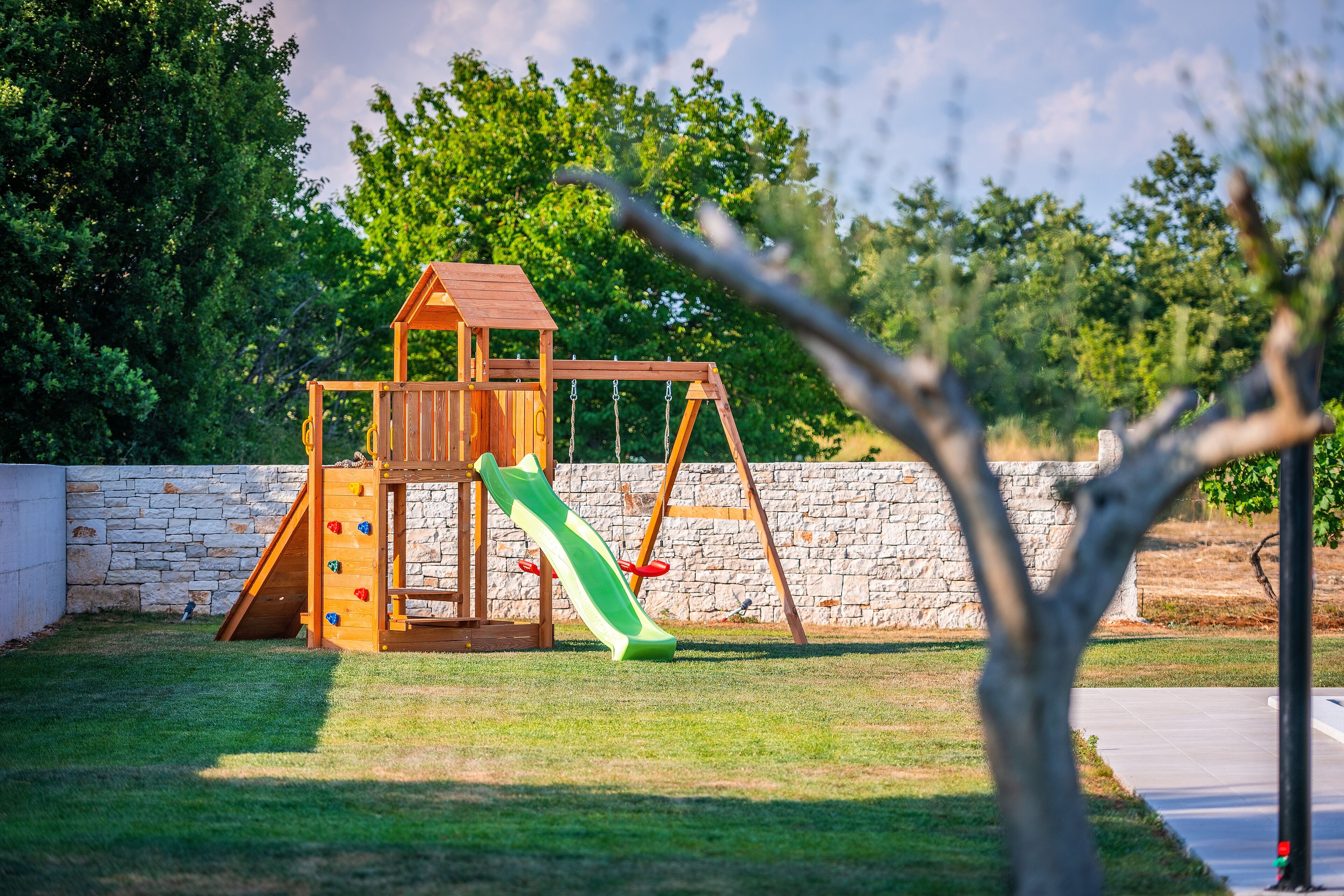 A wooden play structure with a slide and climbing wall, set against a stone wall and surrounded by grass and trees.