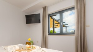Ein Schlafzimmer mit Bett, Wandfernseher, einem Fenster mit Vorhängen und Blick ins Freie.