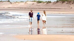 En familj promenerar på en sandstrand med ett lugnt hav och sanddyner i bakgrunden.