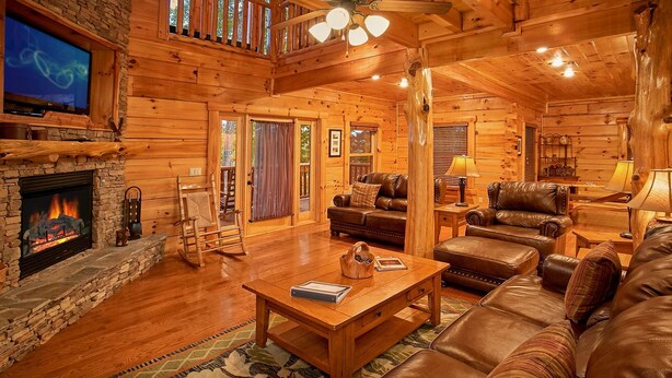 A cozy log cabin interior with a stone fireplace, leather furniture, and a television. - Family Adventure Cabin | Large theater | Views (Sevierville)