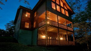 A two-story log cabin with a balcony and large windows, surrounded by trees. - Family Adventure Cabin | Large theater | Views (Sevierville)