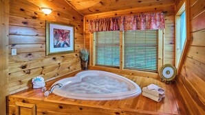 A wooden cabin interior with a hot tub, wooden walls, a window with blinds, and a framed picture on the wall. - Indoor Pool Cabin  | Romantic | Sleeps 4 (Sevierville)