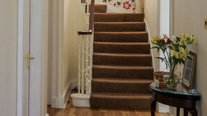 Un escalier avec une rampe en bois et des marches recouvertes de moquette, menant à une pièce dont les murs sont tapissés d’un papier peint à motifs floraux.