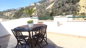 Une table ronde en bois, entourée de chaises, est placée sur une terrasse carrelée. On y voit également un pot de fleurs et, en arrière-plan, une colline parsemée de bâtiments.