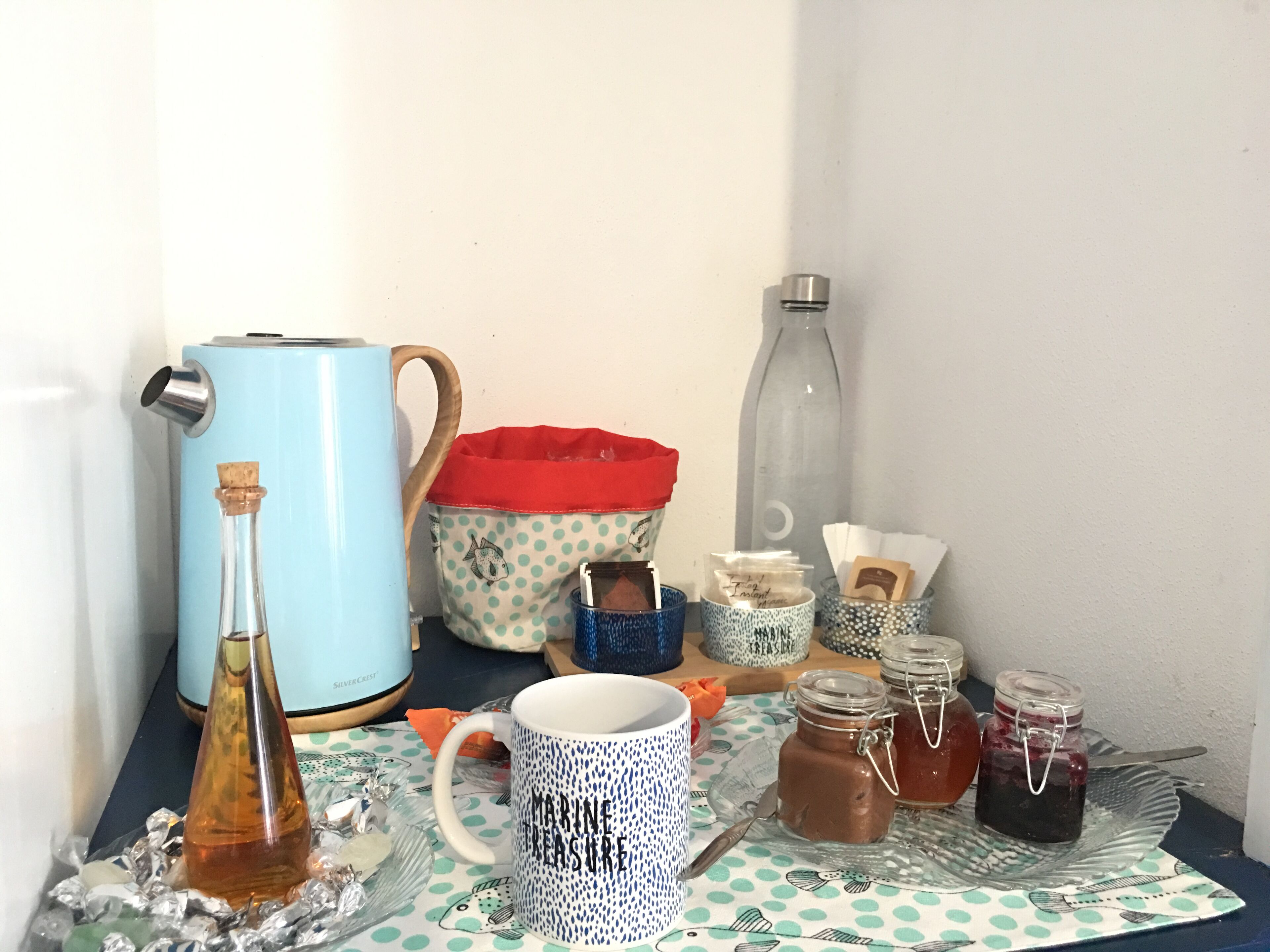 Una mesa con diversos objetos, incluyendo un hervidor azul, un bolso de lunares, frascos de mermelada y una taza con la inscripción 'MARINE PEACE'.