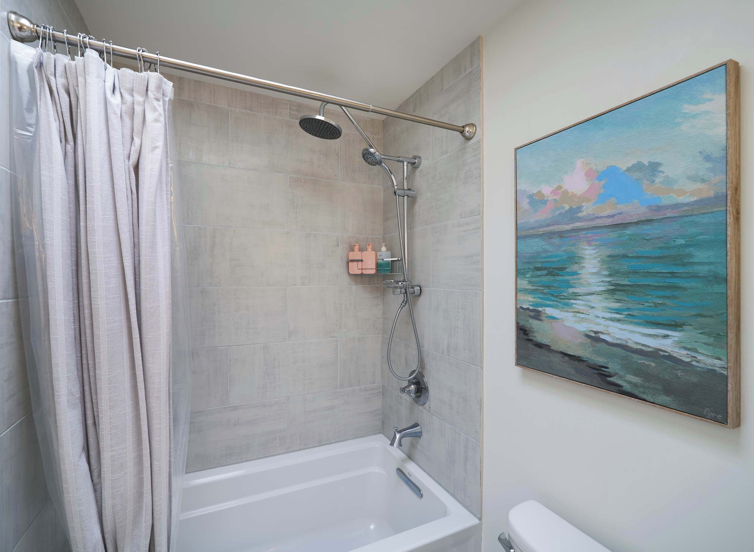 A bathroom with a bathtub, shower, and a painting of a seascape.