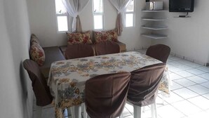 A dining area with a table set for four, a sofa with floral cushions, a TV mounted on the wall, and a window with curtains.