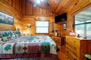 A wooden cabin bedroom with a bed, dresser, TV, and a window with blinds.