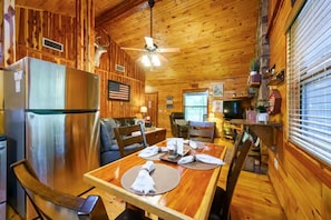 A wooden cabin interior with a dining table, chairs, a refrigerator, a ceiling fan, and a television.