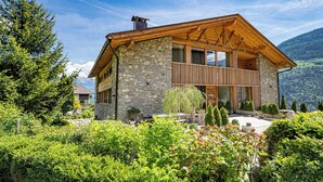 Ein zweistöckiges Steinhaus mit einem hölzernen Balkon, umgeben von üppiger Vegetation und Bergen im Hintergrund.