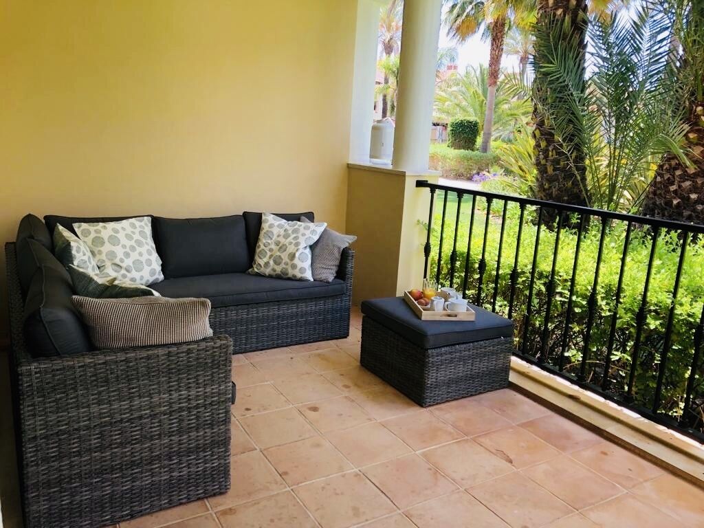 A balcony with a wicker sofa and ottoman, a small table with fruit, and a view of a garden with palm trees.